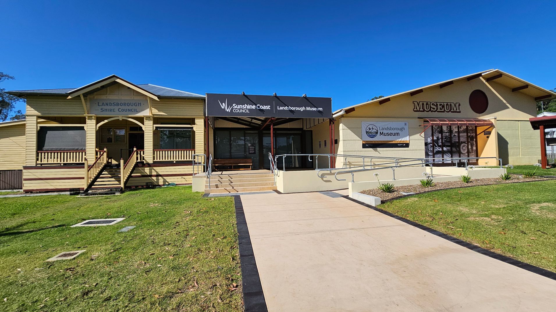 A building with two sections. On the left, a yellow wooden structure with a sign reading "Landsborough Shire Council" above a small porch with steps. In the centre, a covered entrance with a sign for "Sunshine Coast Council Landsborough Museum" and a ramp leading up. On the right, a beige building with a large "MUSEUM" sign and a window displaying a "Landsborough Museum" banner. The foreground features a concrete path and a grassy area with small plants.