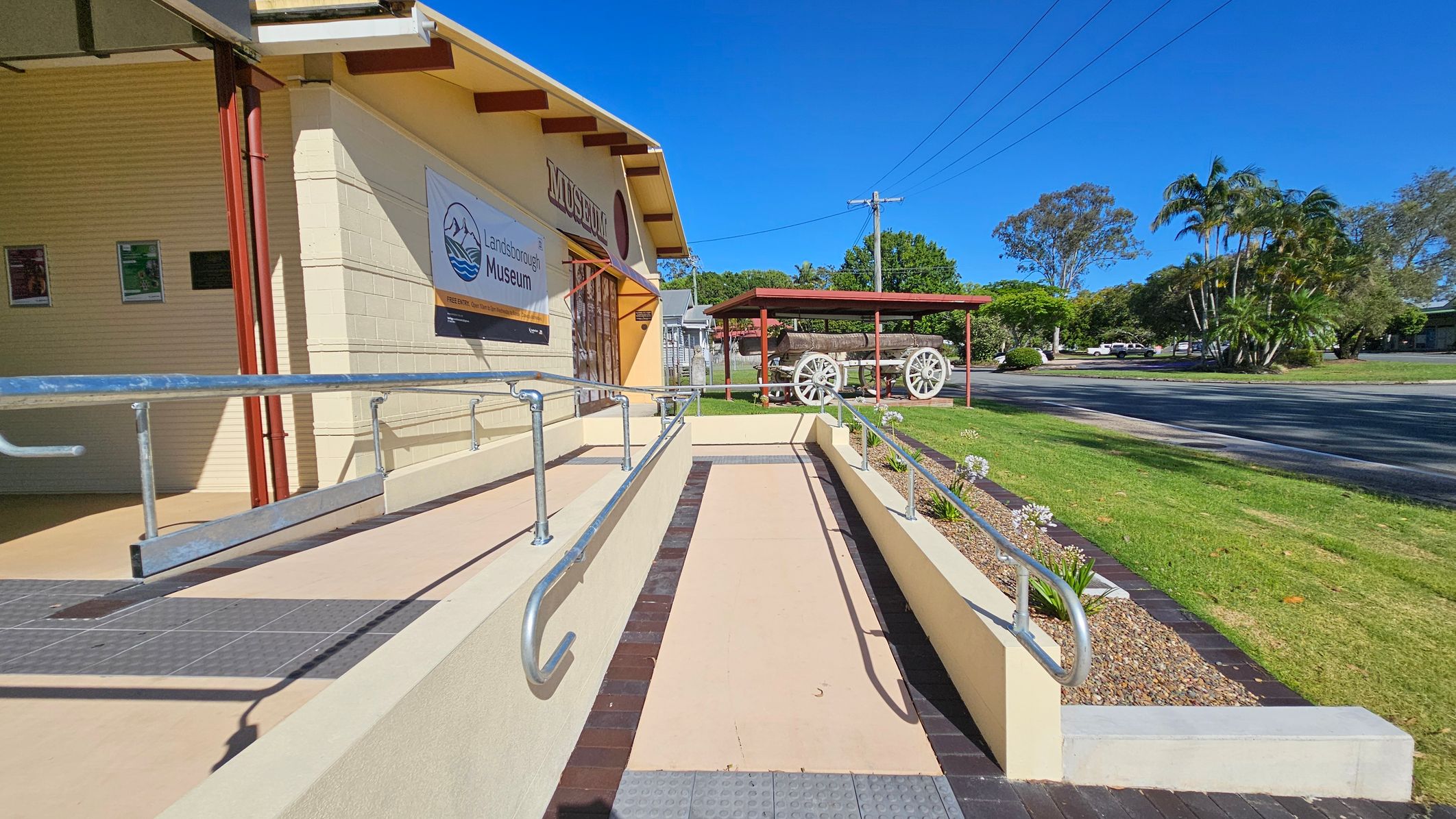 A museum entrance with a ramp and metal railings on the left. The building is light-coloured with a sign reading "Landsborough Museum." Posters are displayed on the wall. On the right, a grassy area with a covered wagon under a red shelter. Trees and a road are visible in the background. The sky is clear and blue.