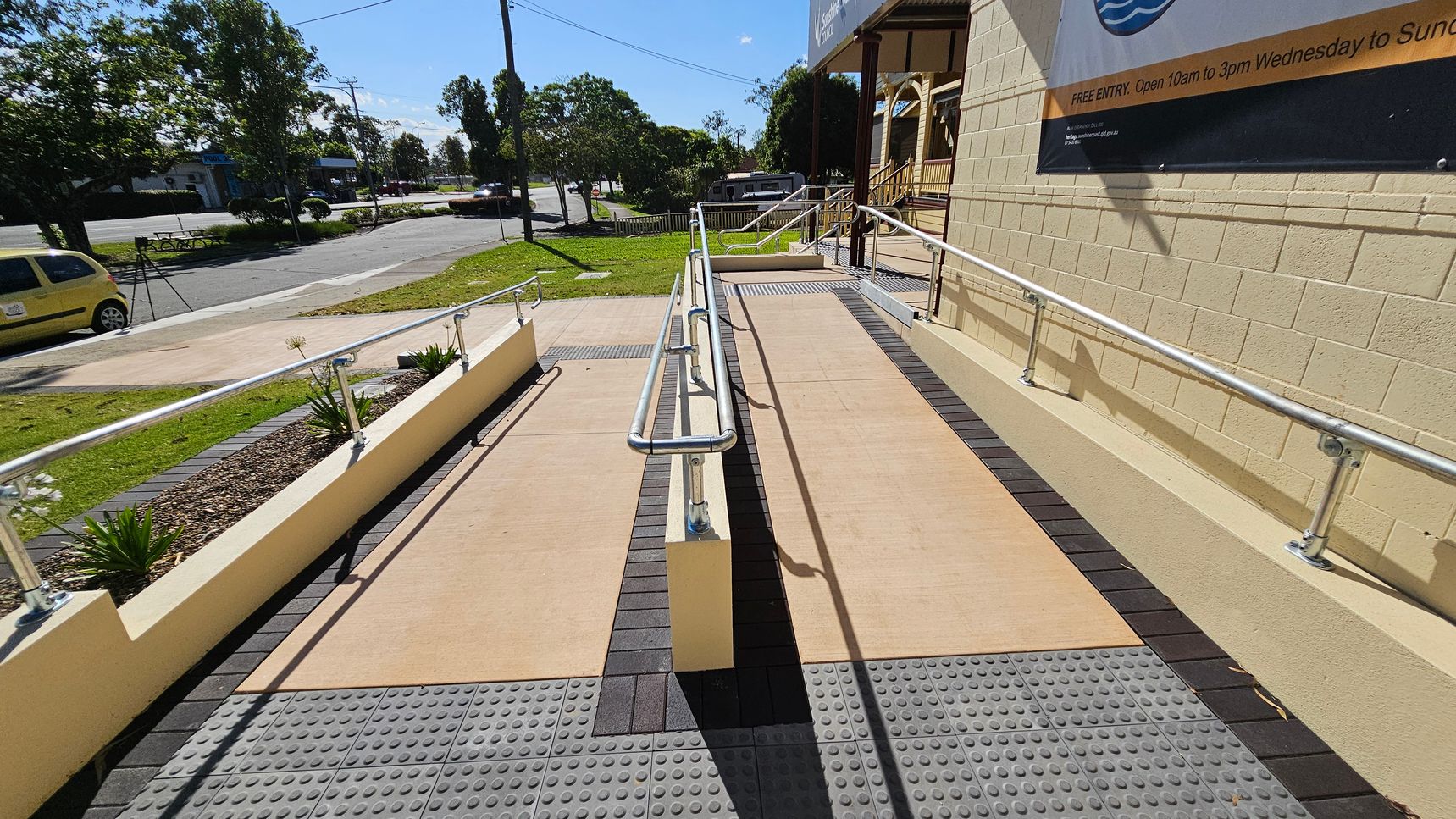 A concrete ramp with metal railings runs alongside a building with a beige brick wall on the right. The ramp is bordered by tactile paving. On the left, there's a small garden area with plants and a yellow car parked on the street. Trees and a petrol station are visible in the background. A large sign is mounted on the building wall.