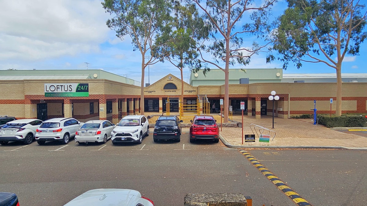 A low-set structure constructed with brown and cream bricks, with the building concourse sharing the brown brick pattern. The facility banner hangs on the left side. The parking area directly adjoins the building, hosting cars of various models and colors. Humps on the road are marked by yellow and black lines. A step-free entrance with a ramp is positioned in the middle. Lamp posts and trees are also visible in front of the building.