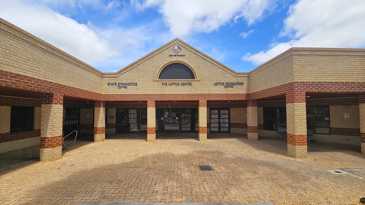 A low-set structure constructed with brown and cream bricks. The roof has a pitched design displaying the city and the facility's logo. The spacious concourse in the middle features the same brown brick pattern and includes a water drainage system. A stainless ramp is visible on the left. Straight ahead is the glass door entrance.