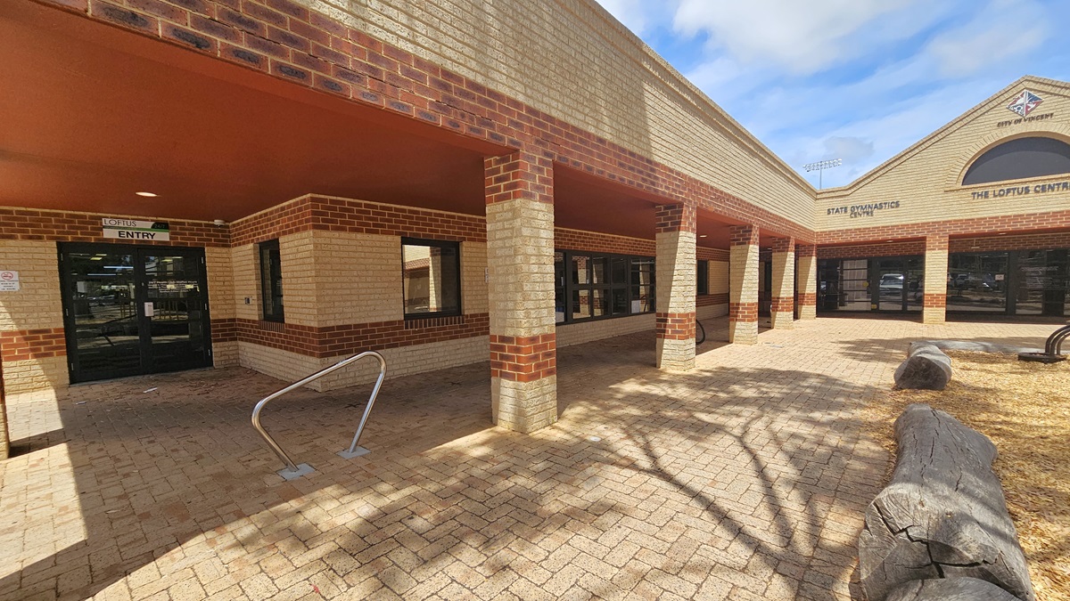 A low-set structure made of brown and cream bricks, featuring a pitched roof displaying the city and the facility's logo. To the left, a glass door marked "Entry" includes a stainless handrail. On the right, a large wooden structure borders the park.