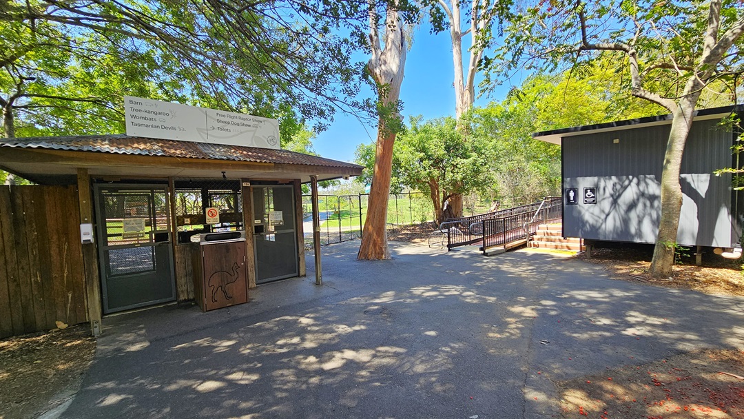 To the left is a shaded structure with a double steel gate. In the middle of the two gates is a wooden rubbish bin featuring an engraved Kangaroo image, with a safety sticker on top. A banner bearing signage is affixed to the roof. To the right is an accessible bathroom with a corrugated wall, accessible via steps and a ramp entry equipped with handrails. Trees surround the area, and the floor is uneven.