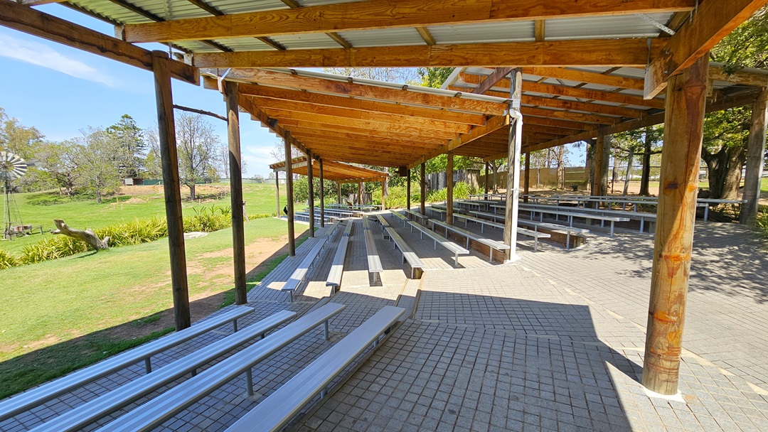 On the left, a landscaped area features green grass, trees, and a wooden windmill structure. Adjacent is a shaded facility with corrugated roofing, wooden beams, and pillars. Beneath, tiered seating for animal show spectators is made of concrete bricks. The surroundings are enveloped by trees.