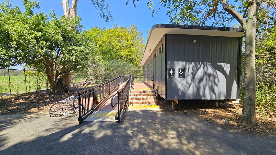 On the right, there's an accessible toilet block featuring grey corrugated walls. Stairs and a ramp with railings, both equipped with yellow truncated domes, are situated on the left. A signage indicating accessibility and gender access is mounted on the wall. Adjacent to the ramp on the left is a dome-shaped metal cage enclosed by a chain metal fence. The surroundings are dominated by large, mature trees.