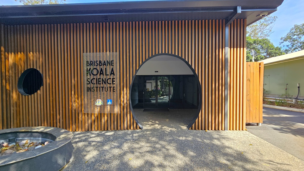 The Brisbane Koala Science Institute. The modern timber structure features round doors and windows. A prominent glass sign that reads “Brisbane Koala Science Institute” is situated on the left side of the entrance. A functional CCTV is visible in the ceiling on the right. A round hole is noticeable on the floor surface to the right.