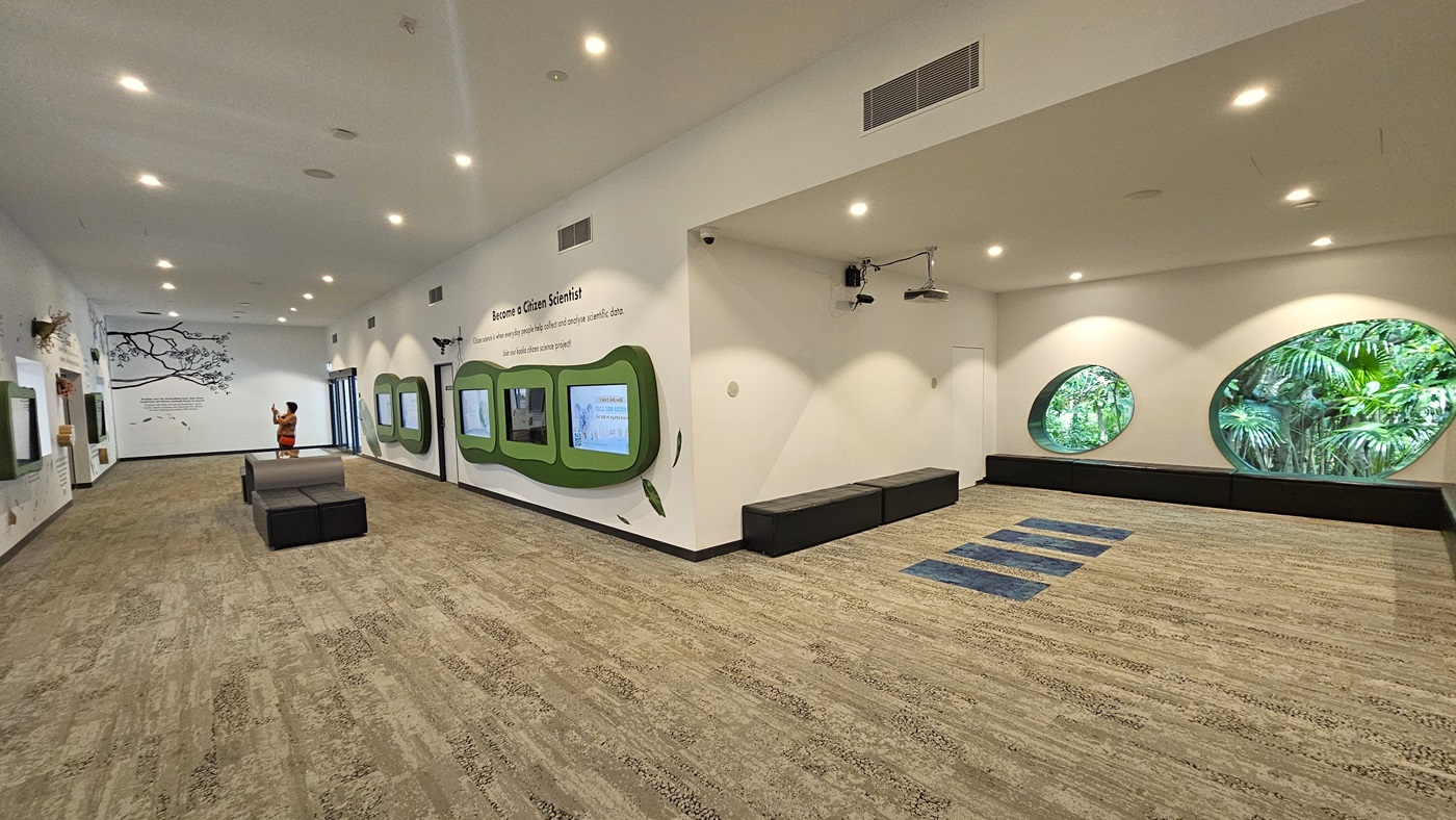 A different view inside the facility. An individual behind the bench seating on the left is visibly taking photos. Another bench seating is situated on the right, beneath the functional CCTV. Fluorescent lighting prominently illuminates the area. Two round holes are apparent on the right side.