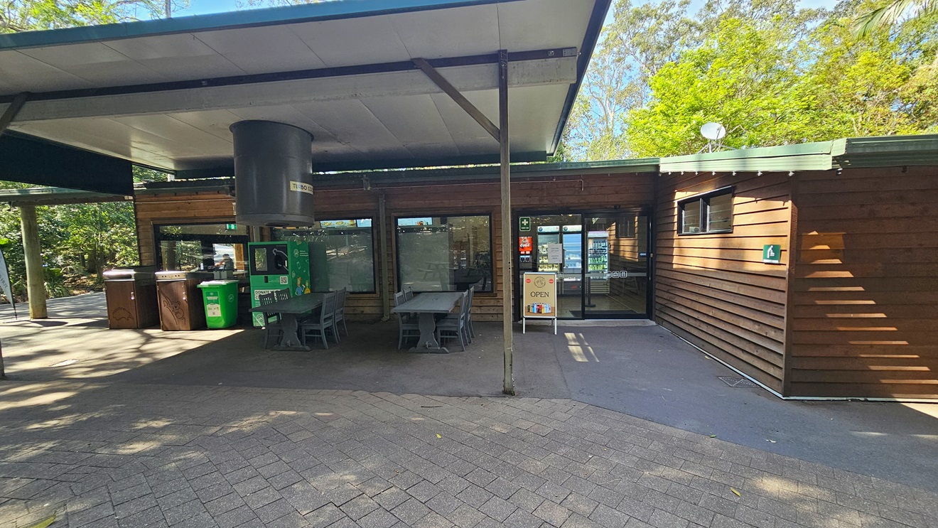 Closer look at the Experiences Hut. A white teardrop flag banner with the facility’s name is visible on the left side. Near the brown timber structure, a shaded verandah with a green directional sign that indicates the way leading to the kangaroo feeding is prominently hanging on the left side of the verandah. On the right, another timber structure is prominent.