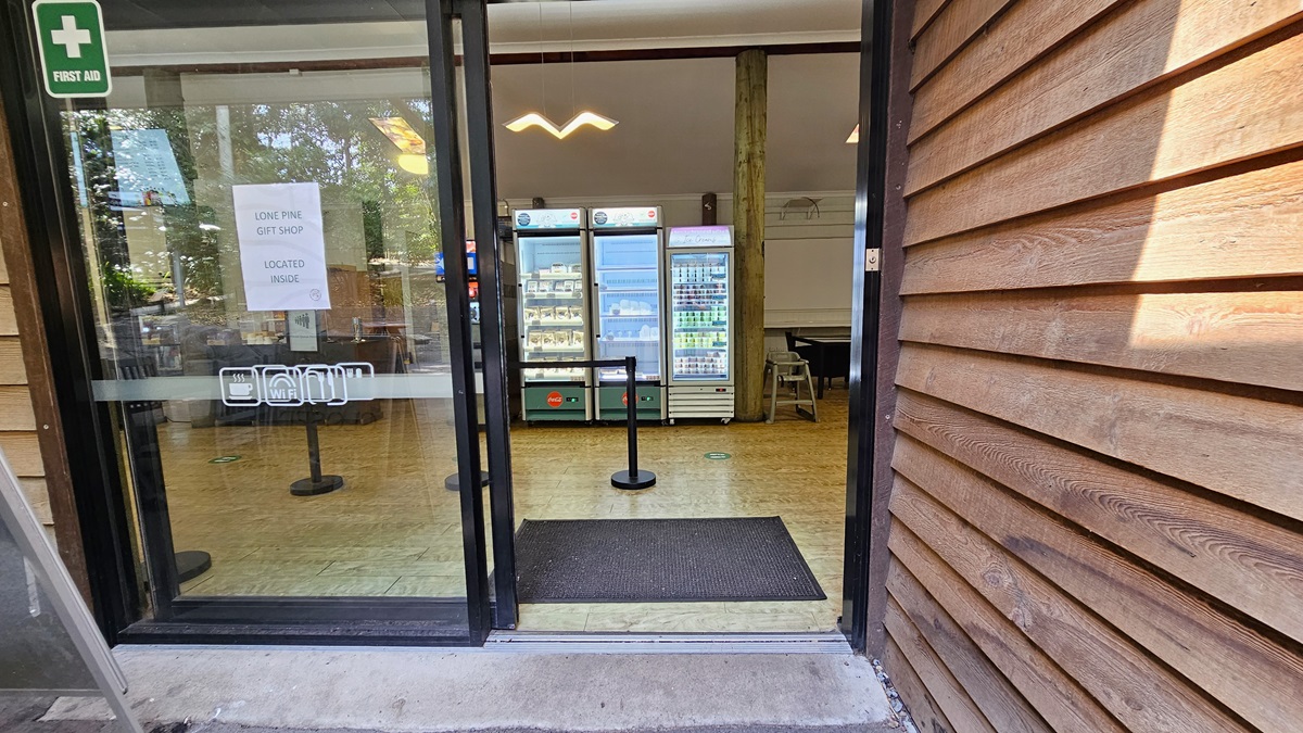 Entryway leading to the inside of the Experiences Hut. On top of the left automatic sliding door, a first aid logo and a sign that indicates that the Lone Pine gift shop is located inside are noticeable. Upon entering, a black floormat and bollards are apparent. The floor surface has timber flooring throughout. Display fridges with snacks are noticeable in the center. The bird-like design of a fluorescent light is prominently hanging on the ceiling.