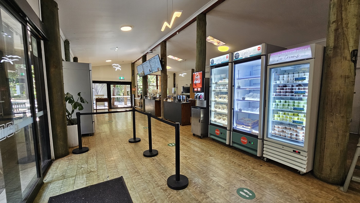 Inside the Experiences Hut. The timber floorings are noticeable in the entirety of the establishment. Three display fridges with a range of snacks are apparent, and next to them is a Coca-Cola freestyle. On the right side of the bollards, the green social distancing logo is apparent on the floor surface. Three flat-screen TVs are noticeable on top of the counter that display the names and prices of the food and beverages.