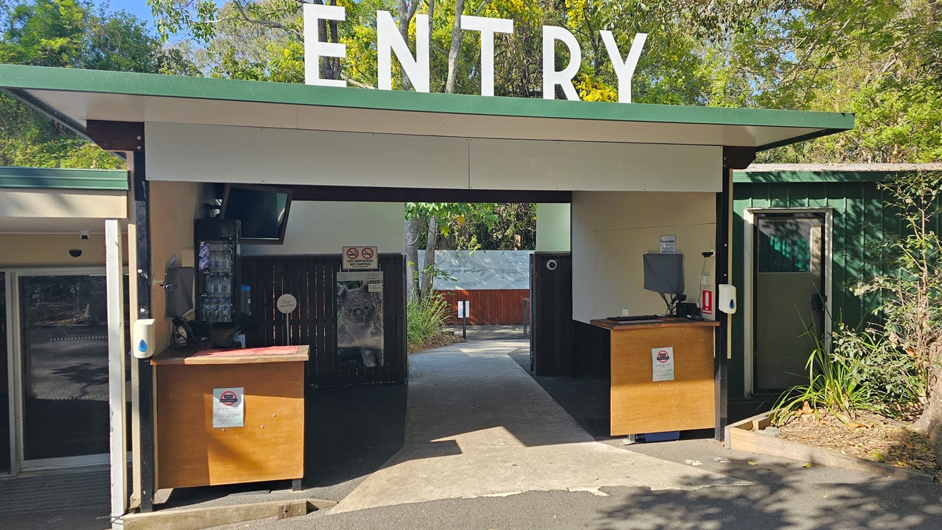 Entryway leading to the ticketing office. A bold “ENTRY” sign is prominent on top of the roof of the cream-structured ticketing office. Two brown desks are apparent with hand sanitizers. Signages are apparent in the entryway. On the ceiling on the left, a TV is apparent, and on the right side is a functional CCTV. Next to the entryway, a green color bond shed is noticeable on the right.
