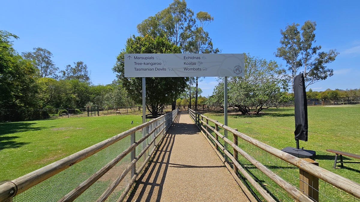 A straight hard gravel surface is flanked by wooden and chain fences on both sides. An arch banner displaying directions is positioned along the path. To the right, a wooden fence and a black banner are present. On both sides, there is green grass and tall trees.