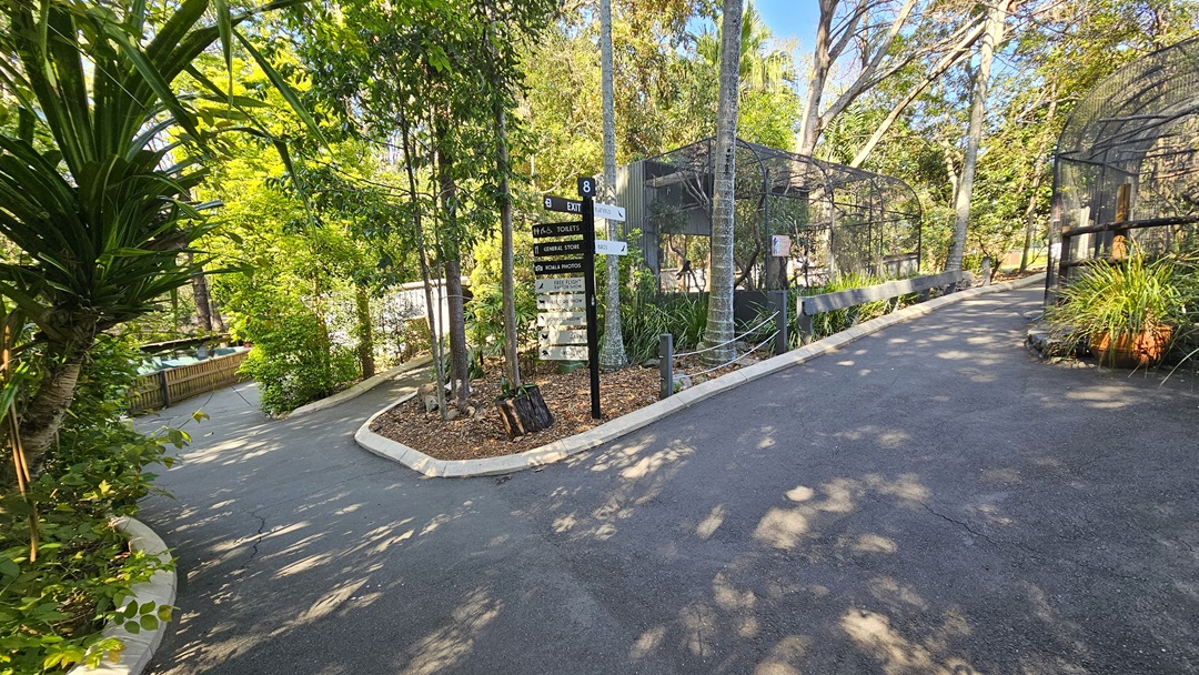 Walkways inside the sanctuary. A large sign that provides information on the attractions in black and white hues is placed in the middle, right after passing the entrance. White concrete garden edges are prominent in the gardens near the walkways. Substantial black timbers are apparent on the right side of the walkway.