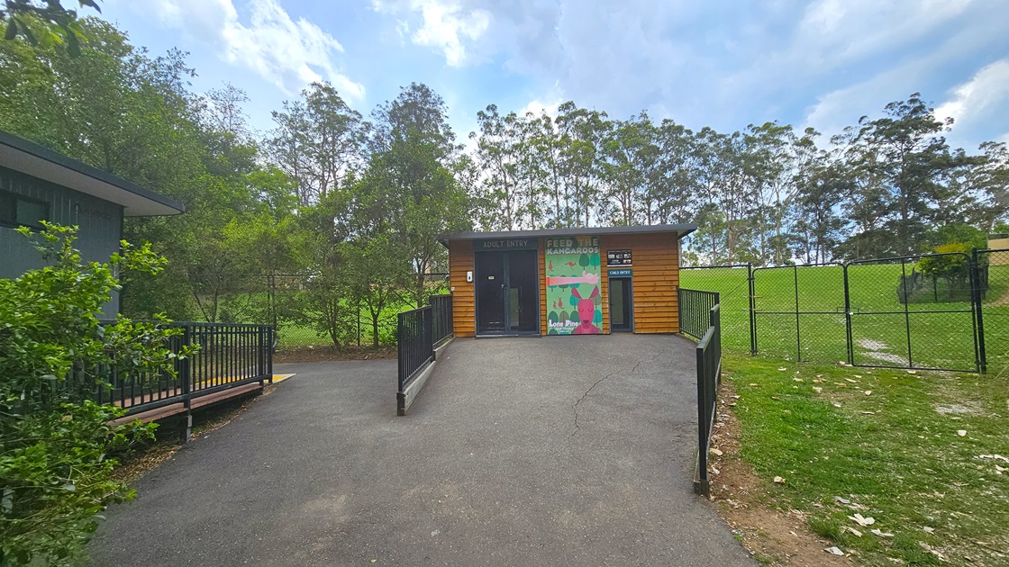 A bitumen entryway leading to the Free Range Feeding. Black hand railings lead to a small timber structure with a poster in the middle that reads “Feed the Kangaroos” is prominent. The timber structure has two entries, with a hand sanitizer equipped on the left entryway. Double-gated structure of the area is apparent on the back.