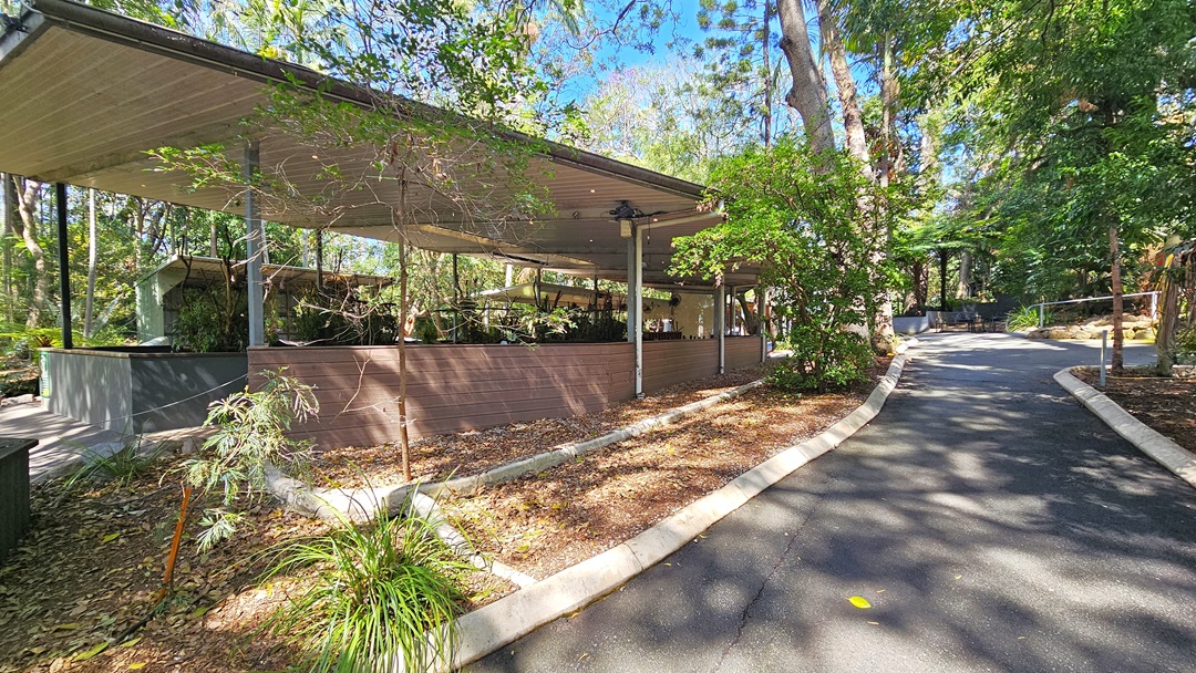 Pathway near a Koala enclosure intended for Koala encounters and photographs. White garden edges are visible throughout the pathways of the facility. Ornaments and tall trees are prominent in the gardens. The Koala enclosure is situated on the left.