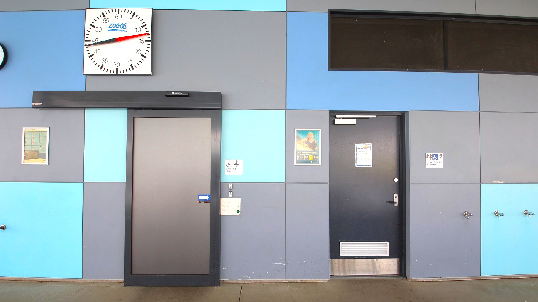 A wall with a geometric pattern of grey and blue panels. On the left, a large clock with "Zoggs" branding is mounted above a grey door. To the right, another door with a poster and a sign indicating accessible changing facilities. Further right, a sign for accessible toilets is visible. The floor is a plain brown surface, and there are small fixtures, possibly taps, on the lower right side of the wall.