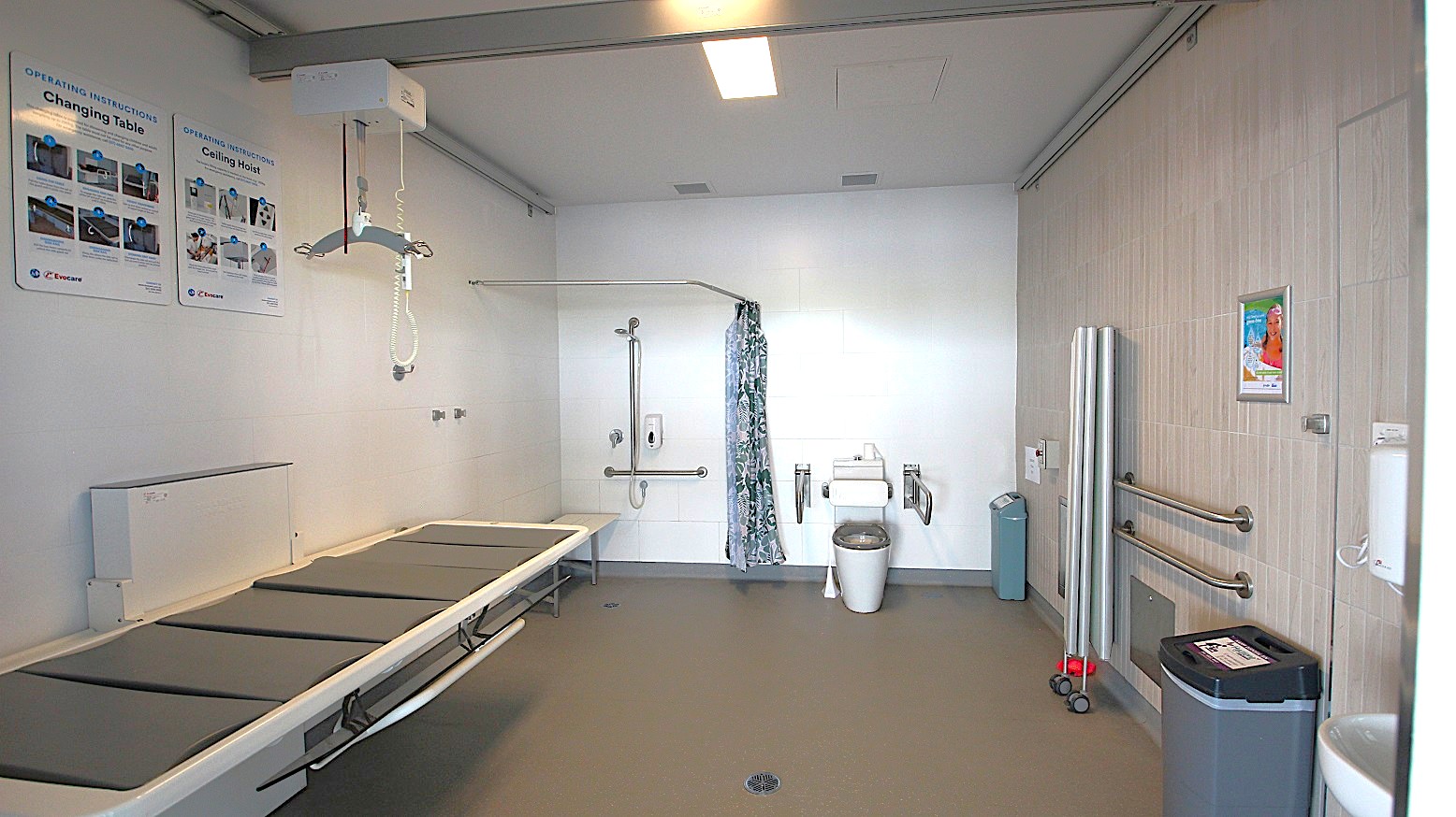 A spacious, accessible bathroom with a grey floor and white walls. On the left, there is a large changing table with padded sections and instructional posters above. In the centre, a ceiling hoist system is installed, and a shower area with a curtain is visible. On the right, there is a toilet with support rails, a bin, and folded equipment against the wall. The ceiling has a rectangular light fixture.