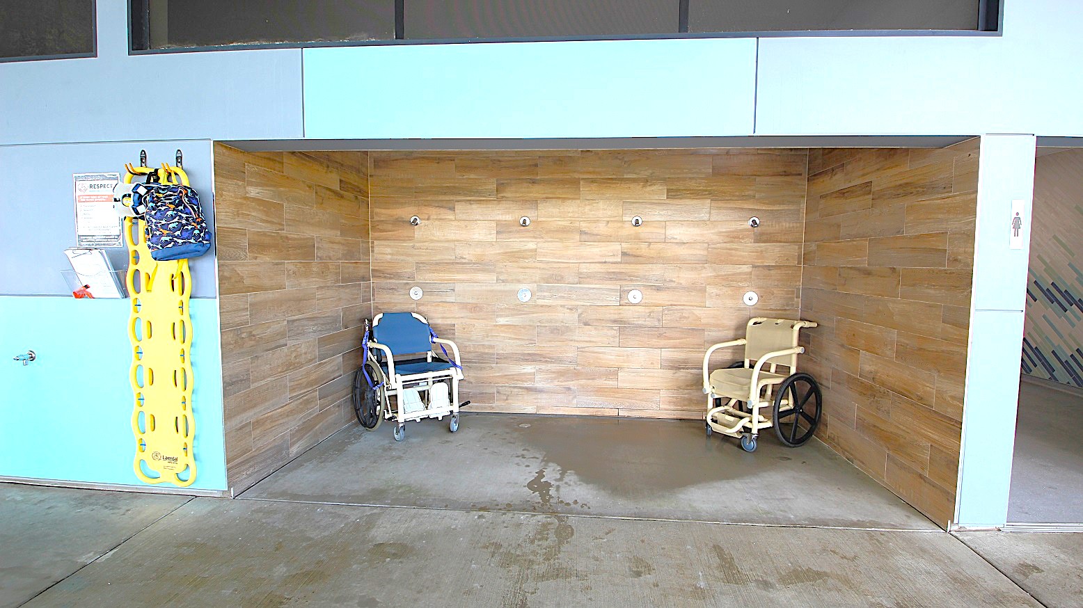 On the left, a yellow spine board and a colourful bag hang on a grey and blue wall, with a sign and a small shelf. In the middle, a wooden-tiled shower area with two shower chairs, one blue and one beige, is visible. The floor is concrete with water stains. The ceiling is not visible.