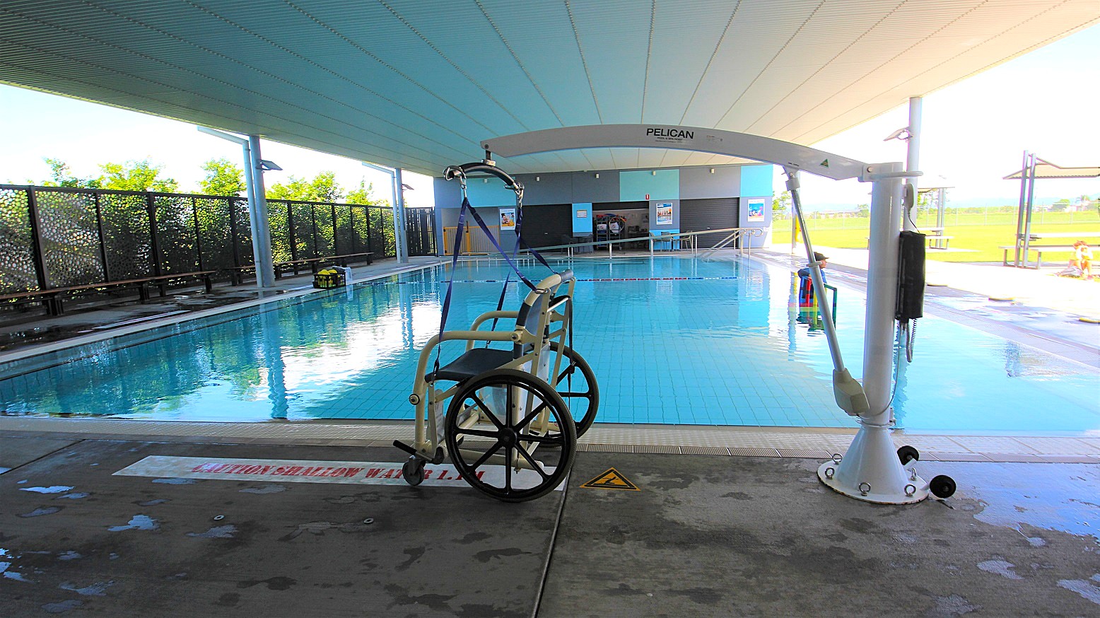 A swimming pool with a hoist system on the right, featuring a wheelchair attached to it. The pool is indoors with a high ceiling and large windows on the left, allowing natural light to enter. The floor is wet and has a caution sign. In the background, there are benches and a fence with greenery visible through it. The pool area is spacious and well-lit.