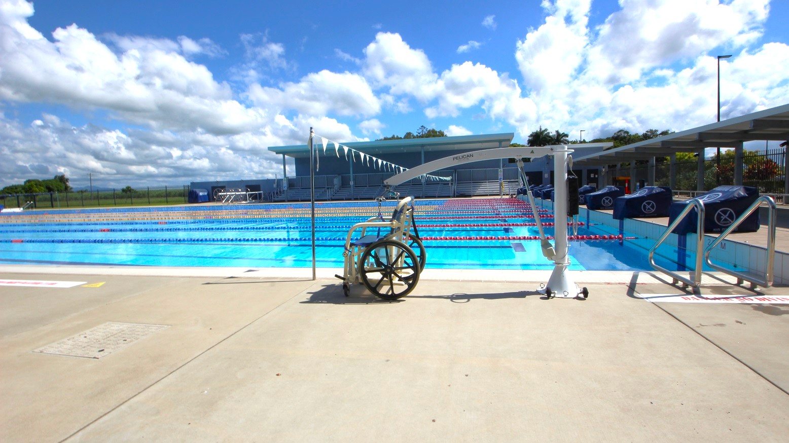 A swimming pool with multiple lanes is in the centre, with lane dividers visible. In the foreground, a wheelchair lift is positioned on the pool deck, next to a wheelchair. On the right, there are steps leading into the pool. The background features a building with a shaded area and a row of flags strung above the pool. The ground is a light-coloured concrete surface.