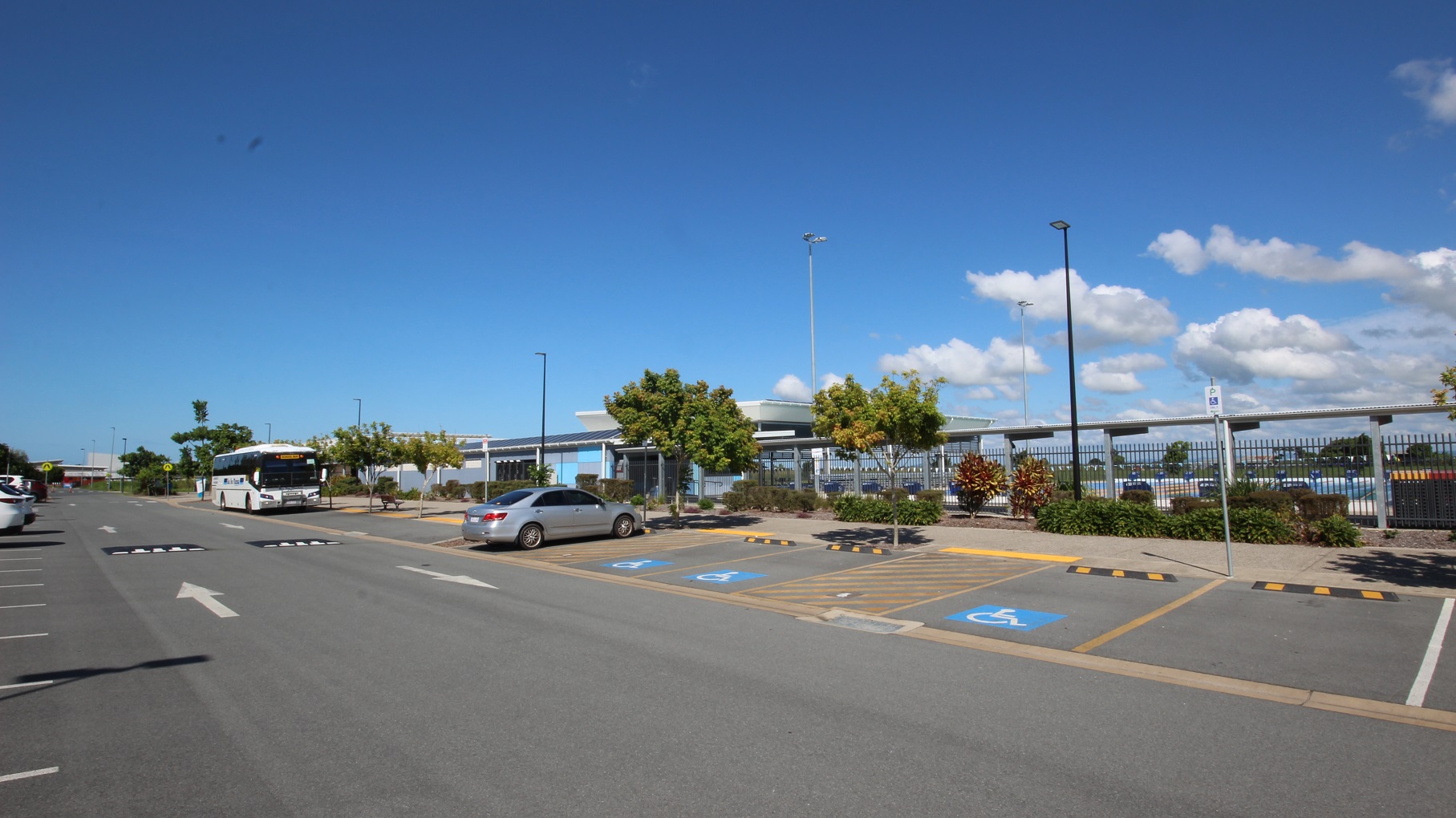 A road with parking on both sides. To the left, cars are parked, occupying several spaces. On the right, accessible parking slots with blue wheelchair signs and yellow diagonal lines are visible. A bus and a grey vehicle are currently parked. Trees and a fence border the right side, and behind is an aquatic centre. Tall street lights are visible.