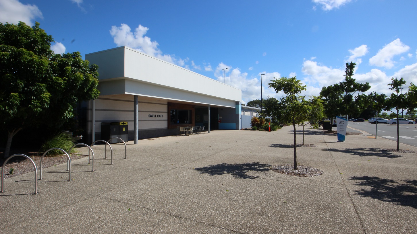On the left is a low-rise facility building with an extended white roof. Tables, chairs, and rubbish bins are under the shade, along with bike racks positioned in front of a tree and grass. To the right, vehicles parked in the car park are visible. Trees line the front and sides, with a retractable banner in place.