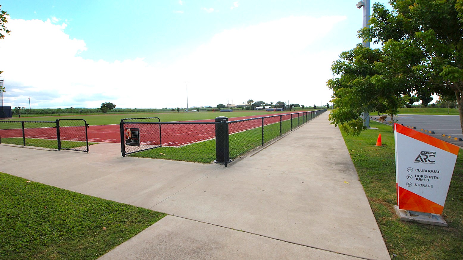 A paved walkway runs alongside a fenced athletic track on the left, with a grassy field inside. On the right, a sign with directional information stands near a row of trees. The path extends into the distance, bordered by a parking area. A single orange cone is visible near the sign.