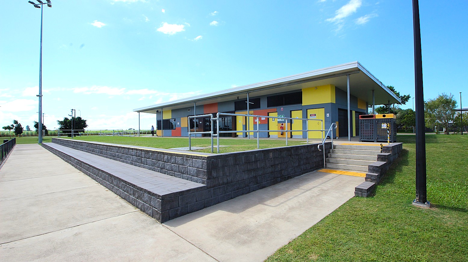 A modern building with a colourful facade featuring yellow, orange, and blue panels. In the foreground, there is a concrete pathway and a low brick wall with steps leading up to the building. Metal railings are present near the steps. The surrounding area is grassy, with a tall streetlight on the left. The sky is clear with a few clouds.
