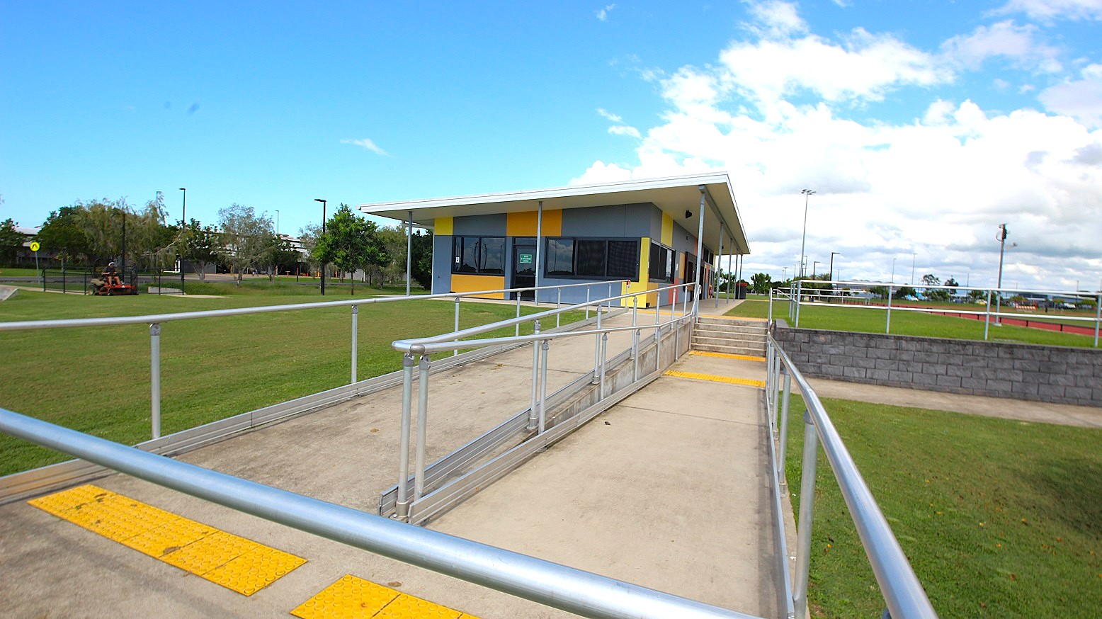 A single-storey building with a sloped roof is in the centre, featuring a colourful facade with blue, yellow, and orange panels. In the foreground, a concrete path with metal railings leads to the building, with a ramp and steps. The surrounding area is grassy, with a few trees and a fence on the left. A low brick wall runs parallel to the path on the right. The sky is partly cloudy.