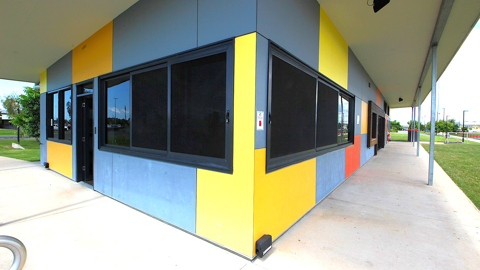 A building with a corner view, featuring a facade of alternating yellow and blue panels. Large, dark-framed windows are set into the walls. The building is situated on a concrete path that extends along its side. The roof is flat and extends outward, supported by metal poles. In the background, there is a grassy area with trees and a fence, and some structures are visible in the distance.