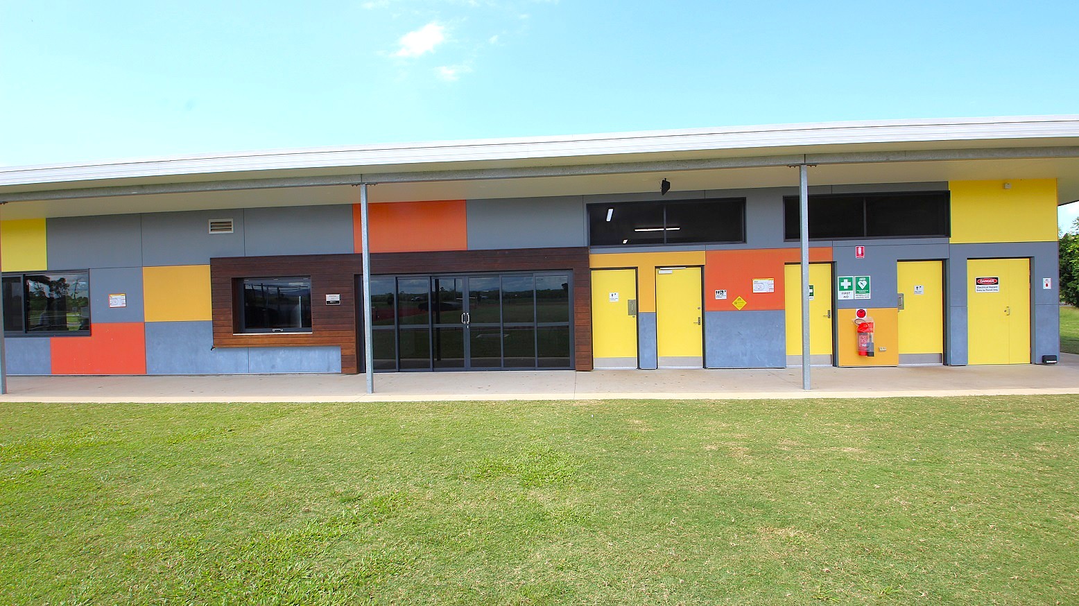 A building with a colourful facade featuring large blocks of yellow, blue, and orange. The structure has several doors and windows, with a prominent set of glass doors in the centre. A fire hydrant is mounted on the wall to the right. The building is supported by metal poles and has a flat roof. In the foreground, there is a well-maintained grassy area.