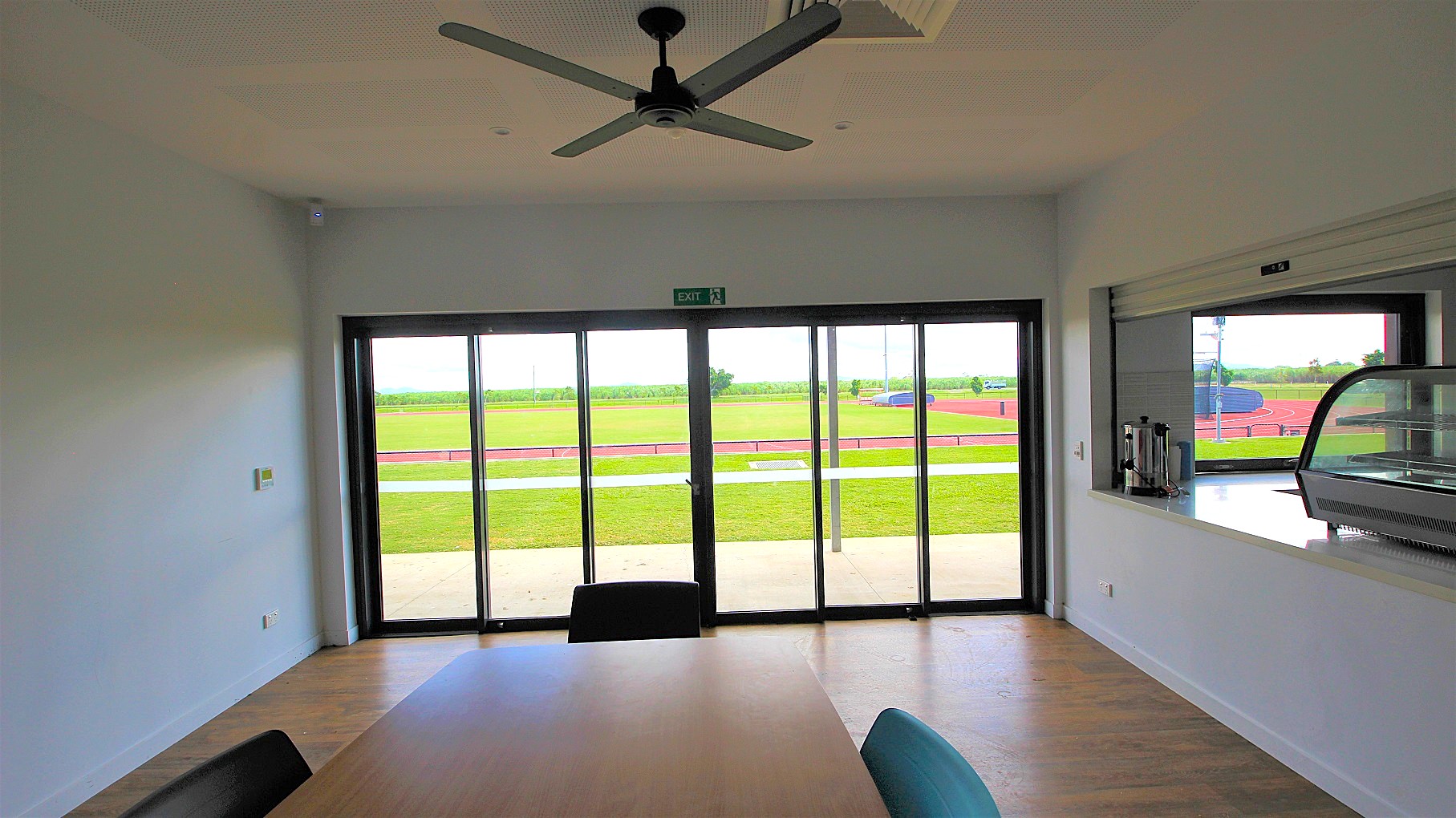 A room with a wooden floor and a ceiling fan. Large glass sliding doors in the centre lead to an outdoor area with a green field and a red running track. On the right, there's a counter with a glass display case and some condiments. A table with chairs is partially visible in the foreground.