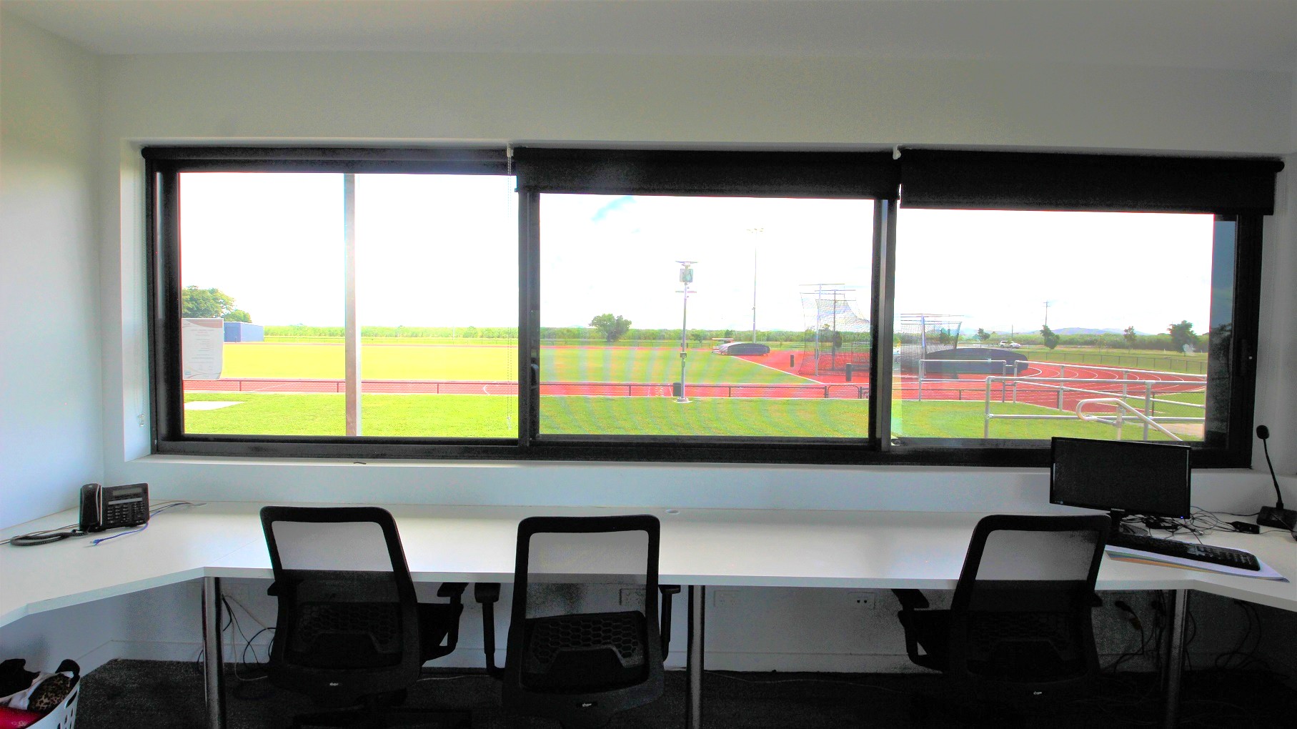 A room with a large window overlooking a sports field with a running track. Inside, there are two black mesh office chairs at a white corner desk. On the desk, a small radio is on the left, and a computer monitor with a keyboard and mouse is on the right. The floor is carpeted in dark grey, and the ceiling is plain white. The view outside shows green grass, a red track, and some equipment in the distance.