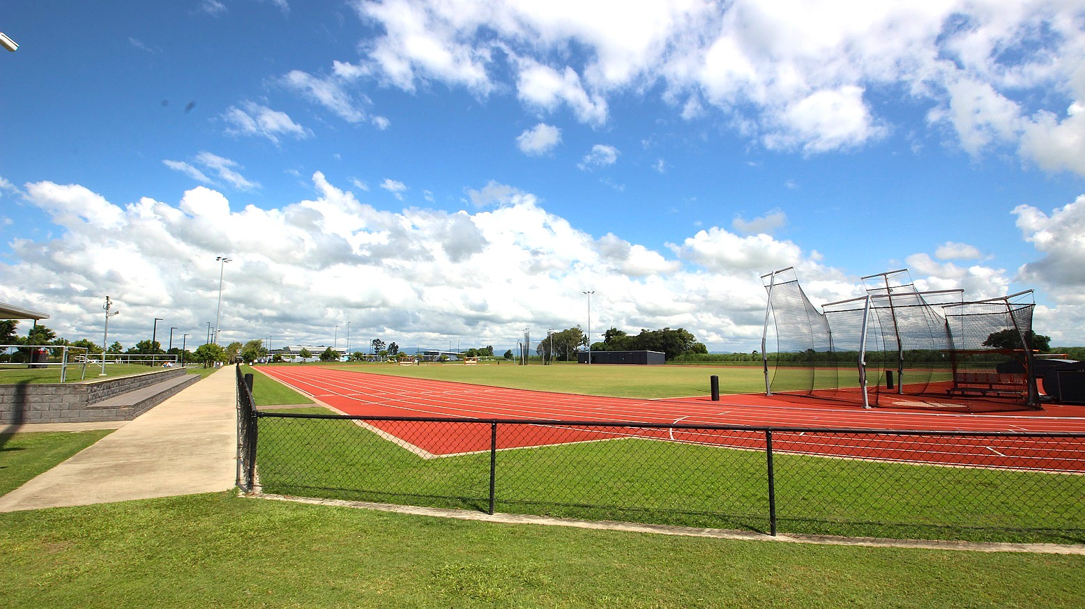 A sports field with a red running track on the right, surrounded by a green grass field. A netted cage structure is visible on the right side of the track. On the left, a concrete pathway runs parallel to the track, with metal railings and a few trees in the distance. The sky is filled with scattered white clouds. A chain-link fence separates the track from the grass in the foreground.