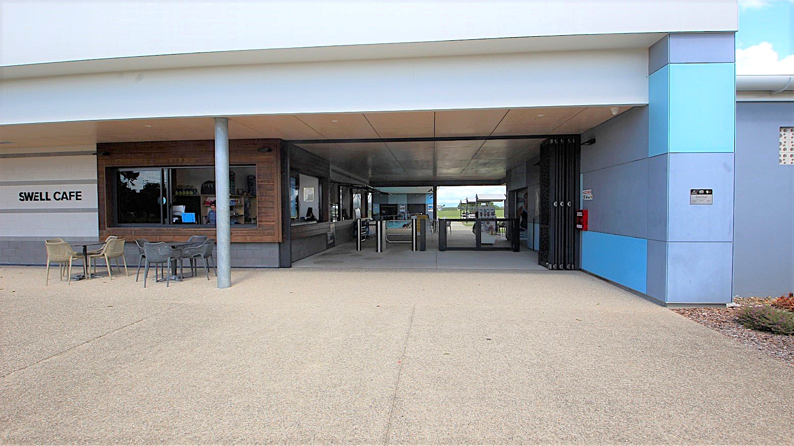 Tables and chairs for exterior dining are placed on a paved area in front of the cafe entrance. A wide serving counter with a glass window is visible. The left internal wall has a timber finish. The cafe name sign is attached to the left side. A ticketed entry turnstile and a folding security shutter are positioned to the right of the doorway. A fire extinguisher is mounted on the external wall alongside the entrance. The concrete exterior is painted white, with blue and grey trim accents.
