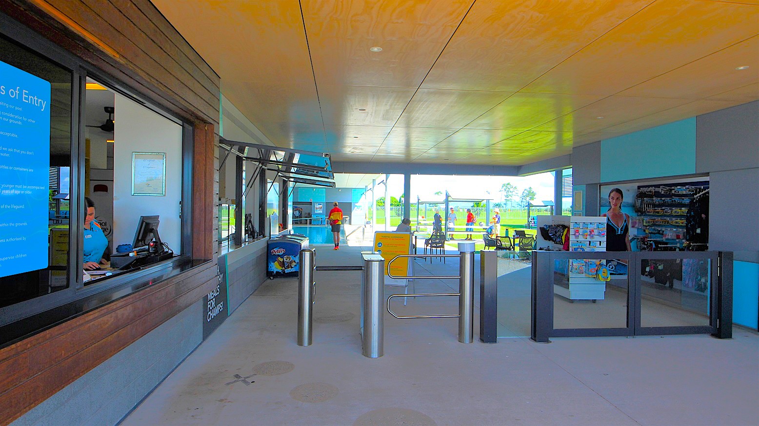 On the left, a reception desk with a wooden front displays a prominent entry condition poster. A staff member stands at a computer. To the right, turnstile gates are visible. Nearby, a small café features an ice cream freezer and a yellow sign. Moveable tables and chairs are arranged. On the right, a retail area showcases swimming goggles and accessories. Further ahead, guests stand under shade structures.