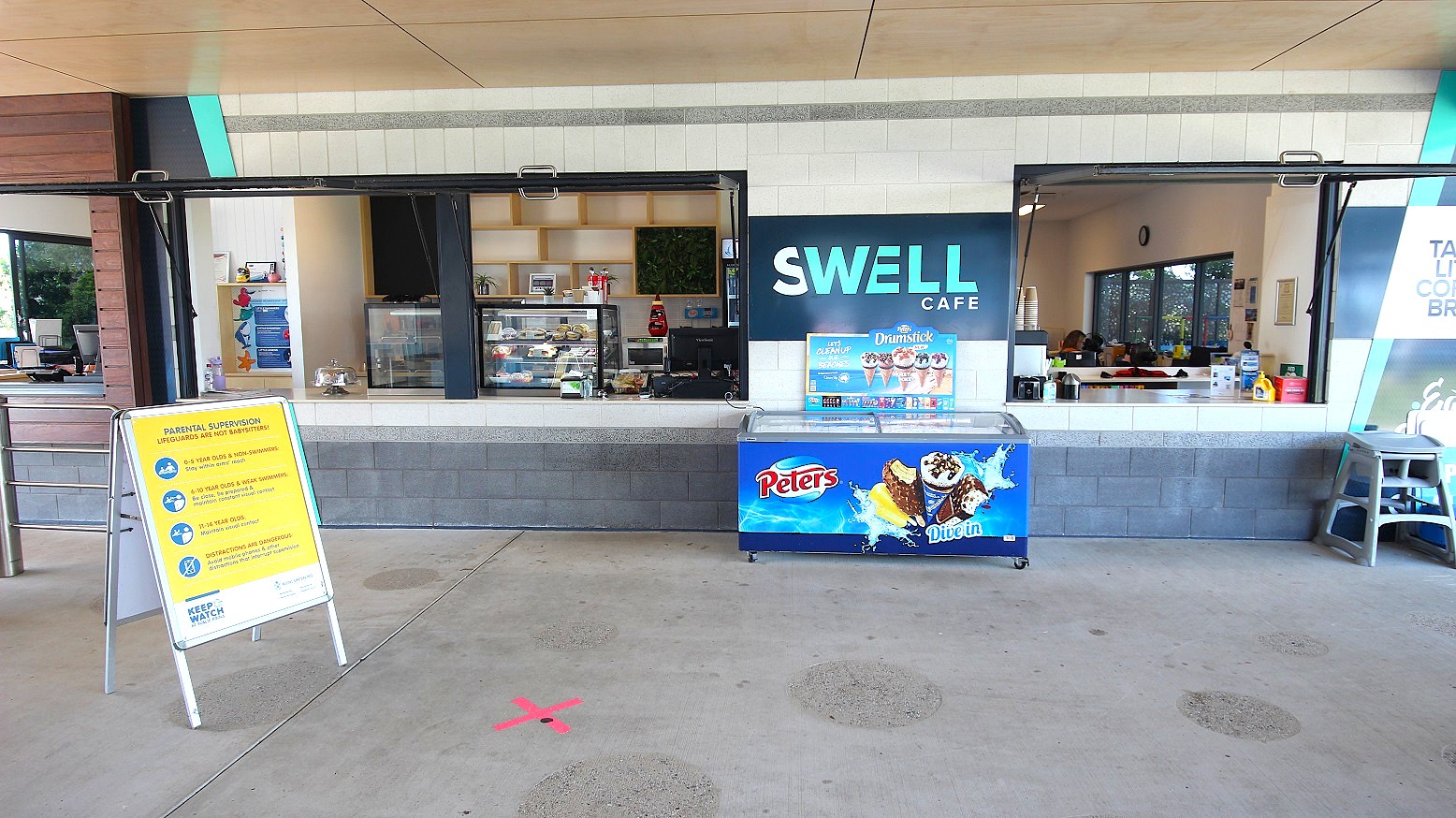 The Swell Cafe displays a grey and white brick facade with a central banner and an ice cream freezer. Inside, food display racks adorn the counter. Wall decorations enhance the ambience. A hot drink dispenser with stacked paper cups is situated on the right. A large banner and a child's high chair are against the wall. A yellow signage is also placed in front.ambi