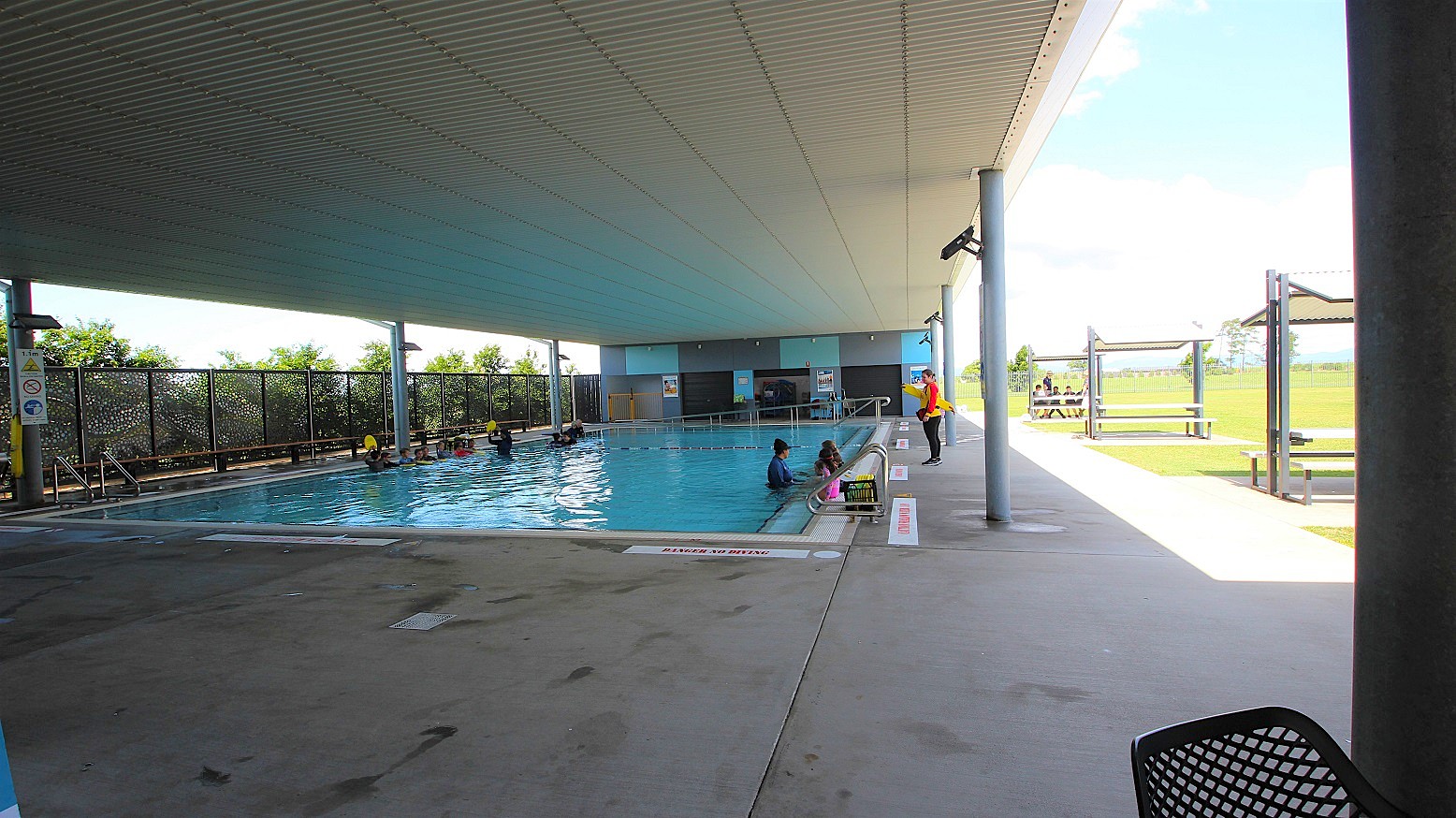 In the centre of an open area lies a pool with a ramp, stairs, and handrails. A metal fence borders the left, accompanied by trees. On the right, shade structures cover picnic tables set on well-maintained green grass. The white corrugated ceiling contrasts with safety banners on the walls. Guests swim while a lifeguard watches over.