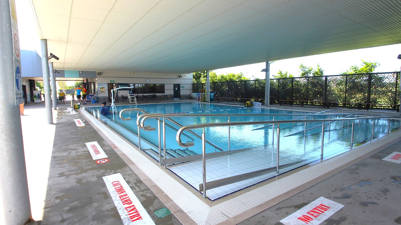 Indoor swimming pool with a ramp leading into the water, surrounded by metal railings. The pool area is covered by a ceiling with a grid pattern. On the left, a walkway with signs on the floor reads "NO ENTRY." In the background, there are people near a seating area and a wall with windows. The right side features a decorative screen with greenery visible through it. The floor is concrete, and the pool is calm and reflective.