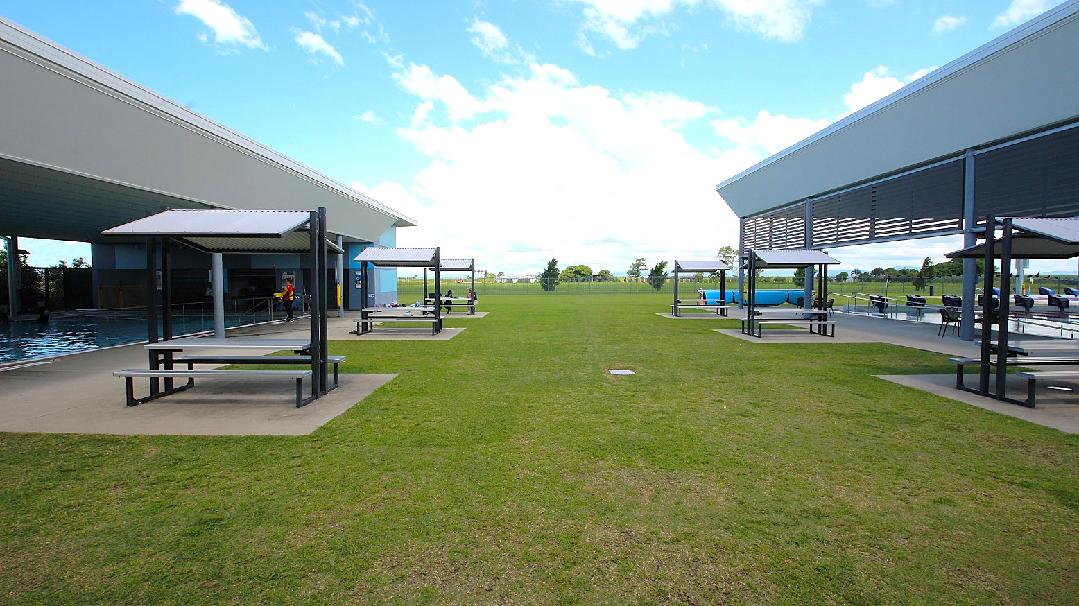On the left, there is a covered area with a swimming pool and a shaded seating area with benches. On the right, another covered area features similar shaded seating. The middle ground is a grassy open space leading to a distant view of trees and a clear sky. The foreground shows a well-maintained lawn, and the structures have modern, angular designs.