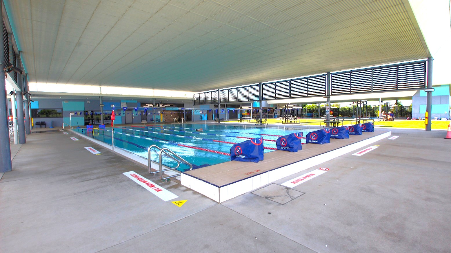 An indoor swimming pool with multiple lanes, separated by red and white lane dividers. Blue starting blocks are positioned at one end. The pool is surrounded by a concrete deck with safety signs. The ceiling is high and white, with slatted panels allowing natural light. On the right, there are tables and chairs on a grassy area visible through large windows. The left side has a walkway with structural columns.
