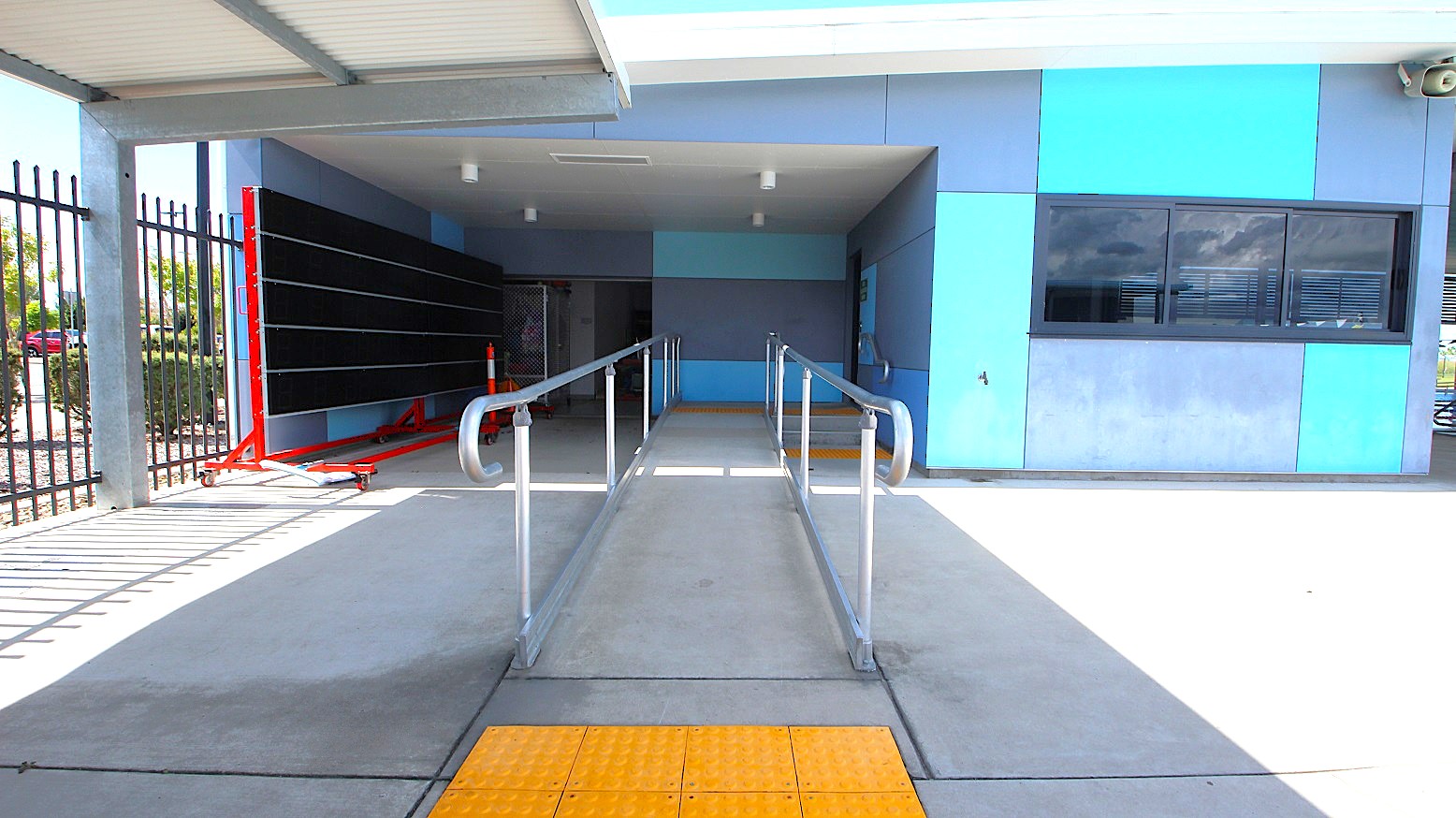 A concrete ramp with metal railings leads into a covered area. On the left, there's a blackboard on a red stand and a black metal fence. On the right, a building with blue and grey panels features a window with closed blinds. The ceiling is flat and grey, with lights installed. The floor has a yellow tactile paving section at the ramp's entrance.