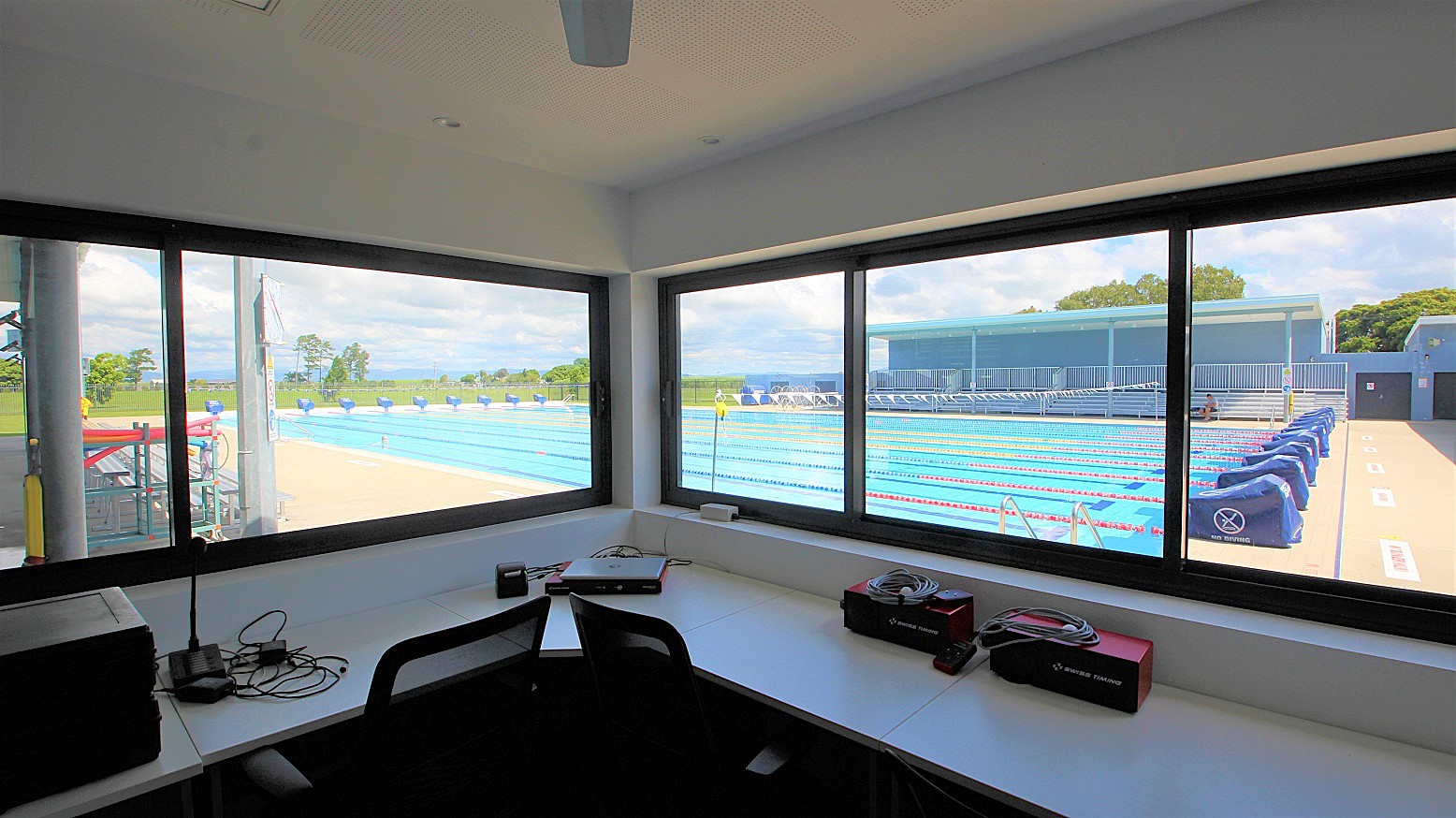 View from inside a room with large windows overlooking an outdoor swimming pool. The pool has multiple lanes with red and blue lane dividers. On the right, there are blue covers on the poolside equipment. The foreground shows a desk with electronic equipment, cables, and a chair. Outside, there is a grassy area and trees in the background. The ceiling is plain, and the floor is not visible.