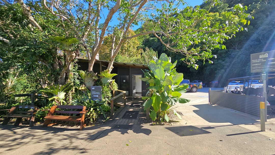 A timber ramp with handrails leads to a low-set building surrounded by trees and gardens on the left. Two wooden benches sit on the concrete driveway. To the right, a paved driveway extends to a parking area with a few golf buggies and vehicles. The backdrop displays a dense forest of trees, with visible signages.