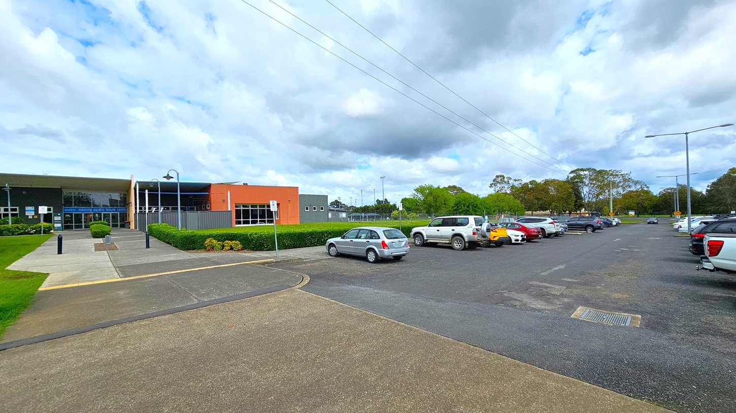 A modern building with large glass windows is on the right, labelled "Aqua Centre" and "Aqua Aerobics". The structure has a slanted roof and is surrounded by a well-maintained grassy area. On the left, there are several trees and a wooden fence running parallel to a concrete path. The path continues on the right, lined with street lamps and more trees. The sky is overcast.