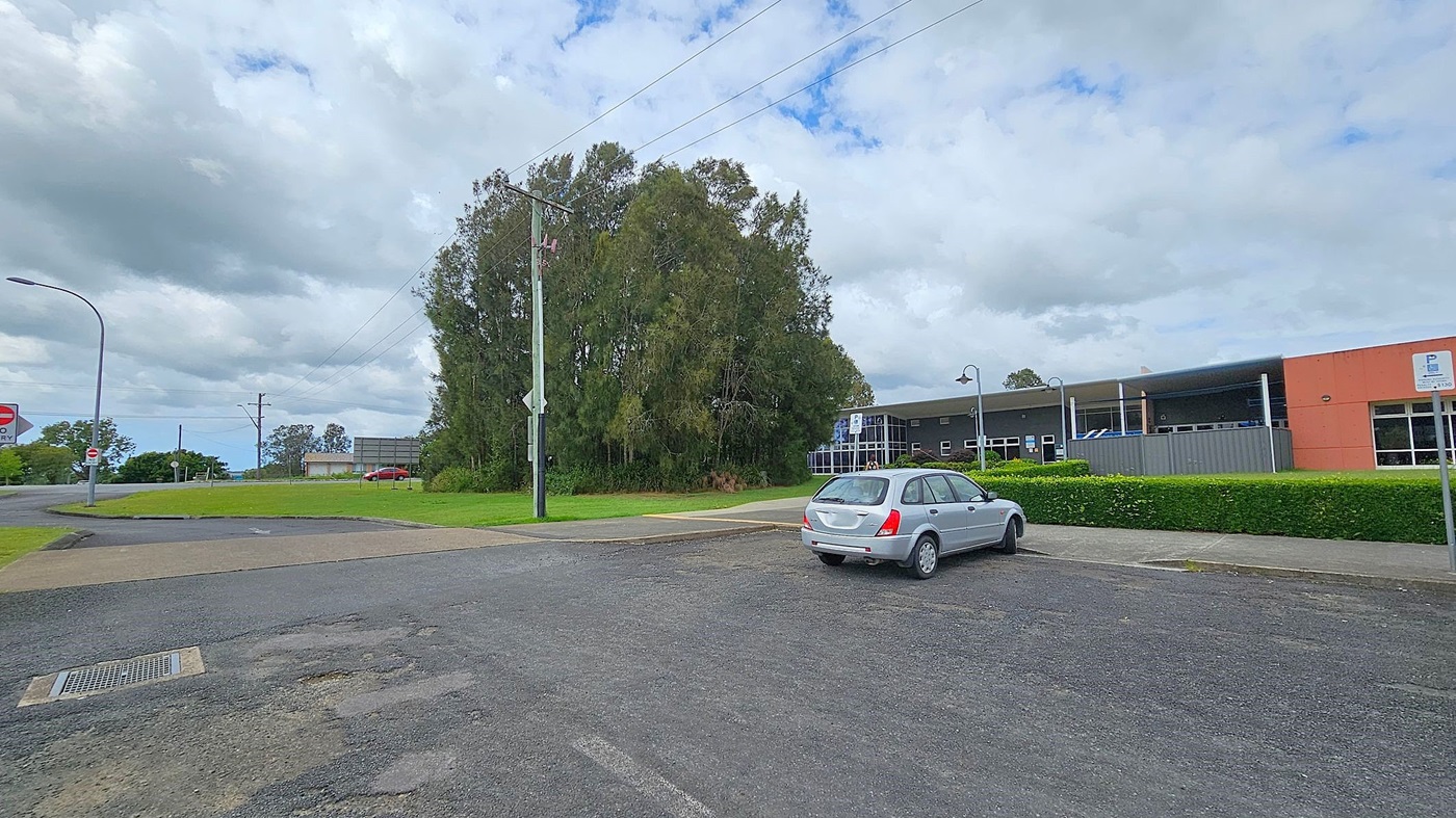 A silver car is parked on a paved area in the foreground. To the right, there's a building with a red and grey facade, surrounded by a neatly trimmed hedge. In the background, a cluster of tall trees stands near a grassy area. A red car is visible in the distance on a road. A streetlamp and a signpost are on the left, with a cloudy sky above.