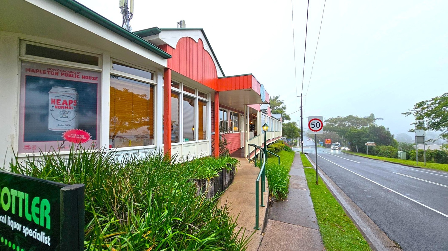 A street scene with a building on the left featuring large windows and a poster advertising a beverage. The building has a red and white facade with a green roof. In front, there is a path lined with green plants and a handrail. On the right, a road with a 50 km/h speed limit sign runs alongside the building. Vehicles are visible in the distance, and trees and greenery line the road. The sky is overcast, creating a misty atmosphere.