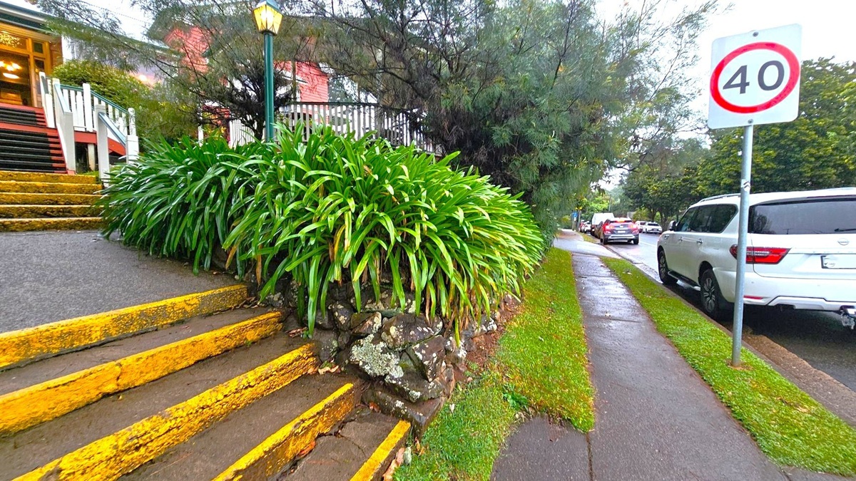 On the left, there are yellow-edged steps leading up to a building with a white railing and a lit lamp post. Dense green foliage lines the path. On the right, a pavement runs alongside a road with parked cars, including a white SUV. A speed limit sign indicating "40" is visible. Trees and bushes are in the background, and the ground appears wet, suggesting recent rain.