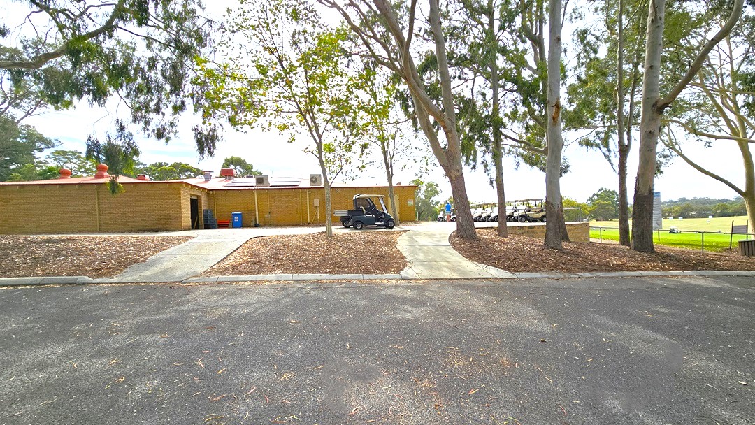 Two concrete pathways ascend, guiding towards a low-rise brown brick building on the left. Adjacent to the building, golf carts are neatly parked in a row on the right, and another is positioned between the two pathways. On the right, the hilly side is populated by tall trees. Furthermore, a spacious green area is enclosed by a steel chain fence on the right.