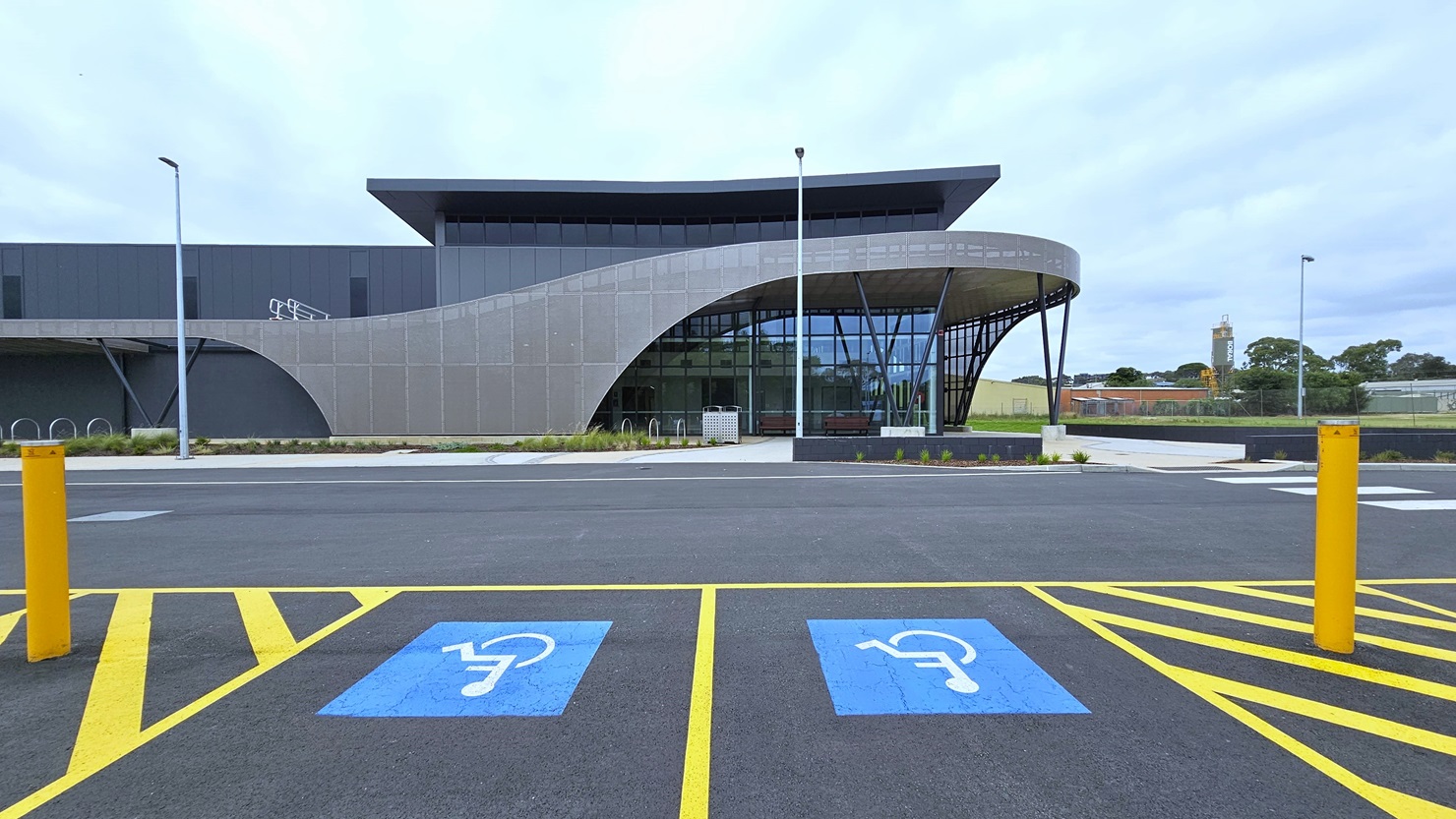 Accessible car parking spaces are marked with signs and yellow bollards. Adjacent is a modern facility building made of concrete, glass, and beams. A grass area lies to the left, bordered by tall lamp posts along the road. Bike racks are positioned at the sides of the building. A concrete plant box hosts plants and small trees.